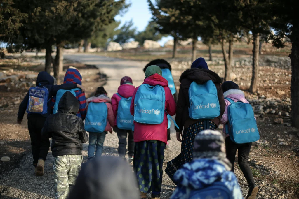 Gruppe af børn med UNICEF-rygsække går ad en sti mod skole eller aktivitetsområde.